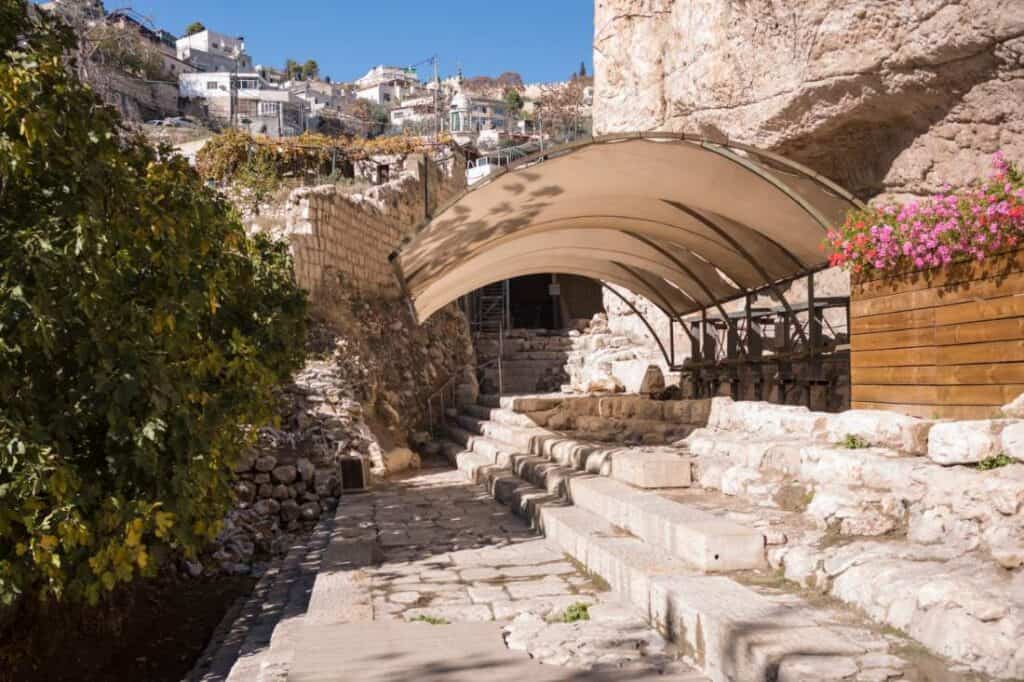 The excavated stepped street and pool area at the Pool of Siloam in Jerusalem, with a canopy over the dig and the houses of Silwan rising behind